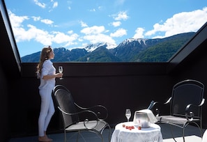 A balcony with a mountain view, two chairs, a table with a bottle and glasses, and a person holding a glass.