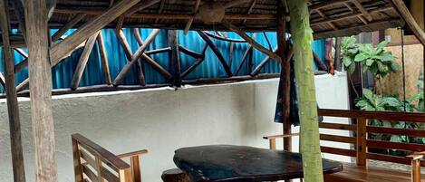 Un espace extérieur aménagé avec des sièges en bois et un toit de chaume, entouré d’un sol carrelé en pierre et d’un mur bleu.