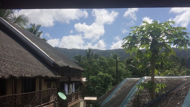 Ein traditionelles Gebäude mit einem Reetdach und einem hölzernen Balkon, umgeben von üppigem Grün und blauem Himmel.