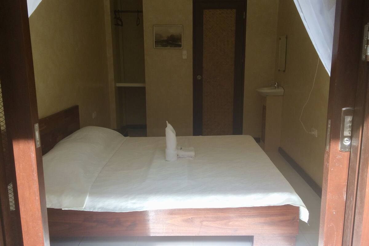 Ein kleiner, schwach beleuchteter Raum mit einem Einzelbett, einer Tür, einem kleinen Waschbecken und einem gerahmten Bild an der Wand.