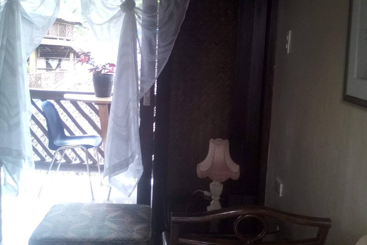Ein Zimmer mit einer Holzbank, einem kleinen Tisch mit Lampe und durchsichtigen Vorhängen.