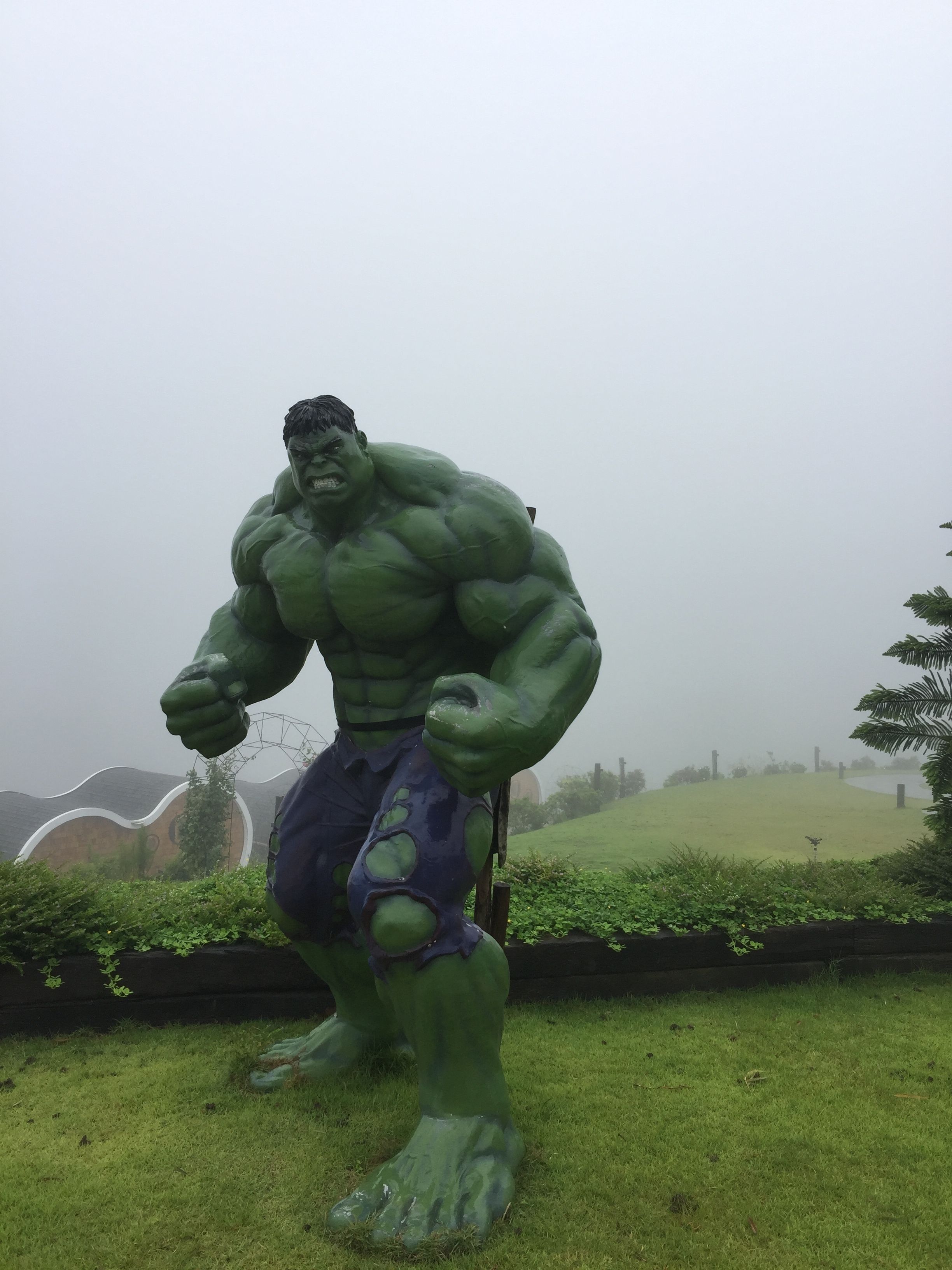 Una gran estatua de Hulk en un área con césped, con colinas y un cielo nublado al fondo.