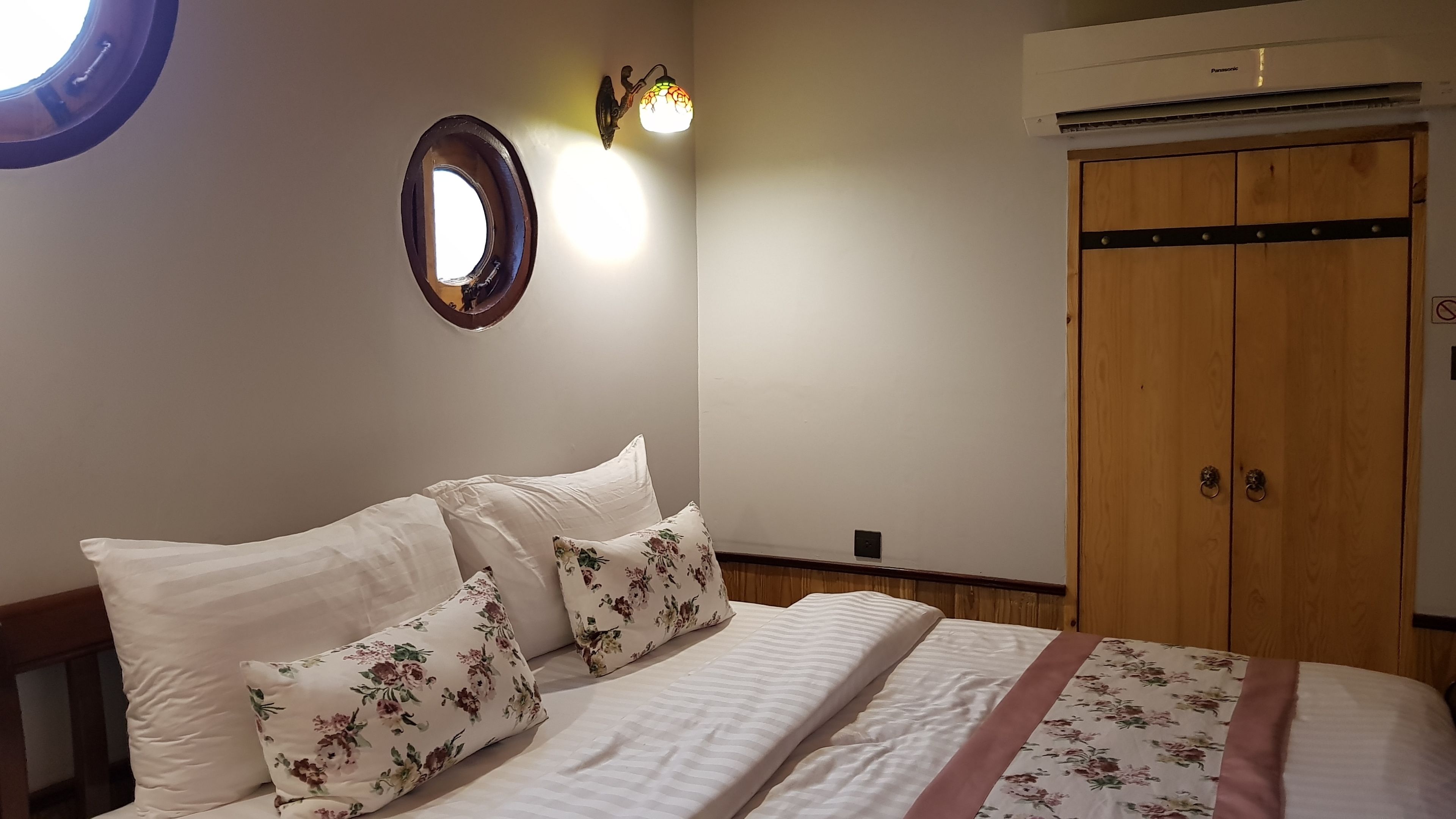 Camera da letto con un letto, cuscini floreali, un punto luce a parete, un'armadio in legno e un condizionatore d'aria.