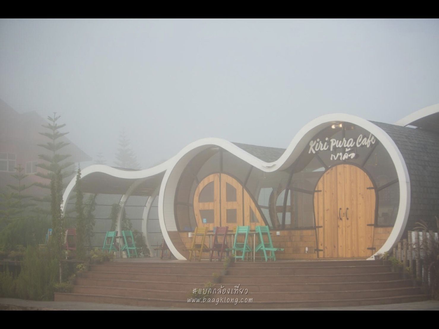 Een modern gebouw met een uniek, gebogen ontwerp, voorzien van een houten deur en een bord waarop 'Kiri Pura Cafe' staat.