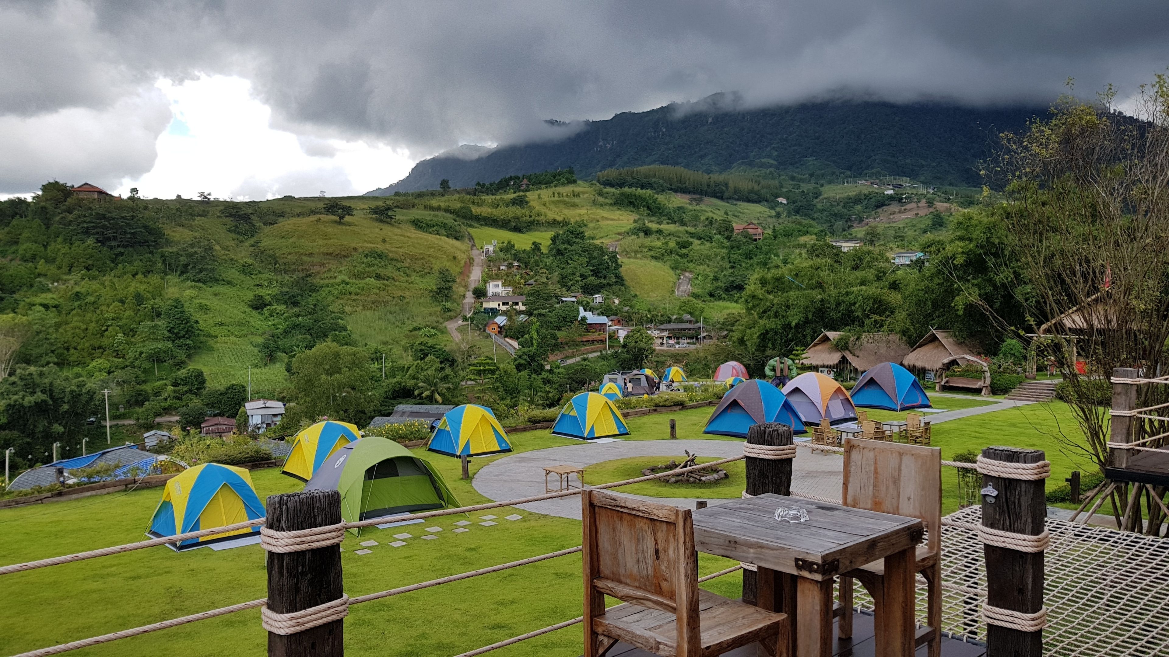 A campsite with multiple tents, wooden tables, and chairs, set against a backdrop of green hills and a cloudy sky.