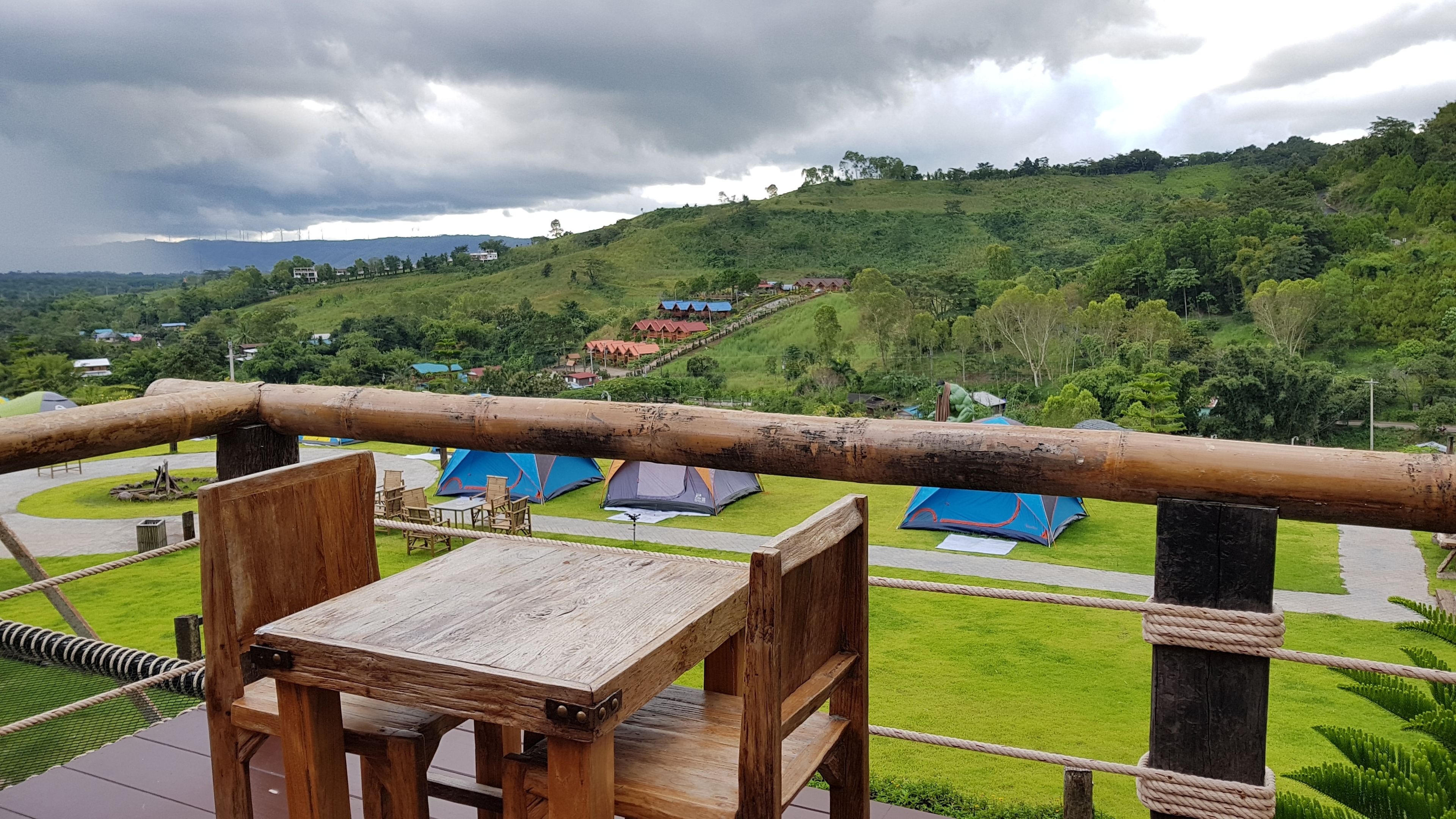 A wooden deck with a table and chairs, overlooking a grassy area with blue tents and houses on a hillside.