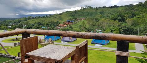 Un terrazzo in legno con tavolo e sedie, affaccia su una zona erbosa con tende blu e case su una collina.