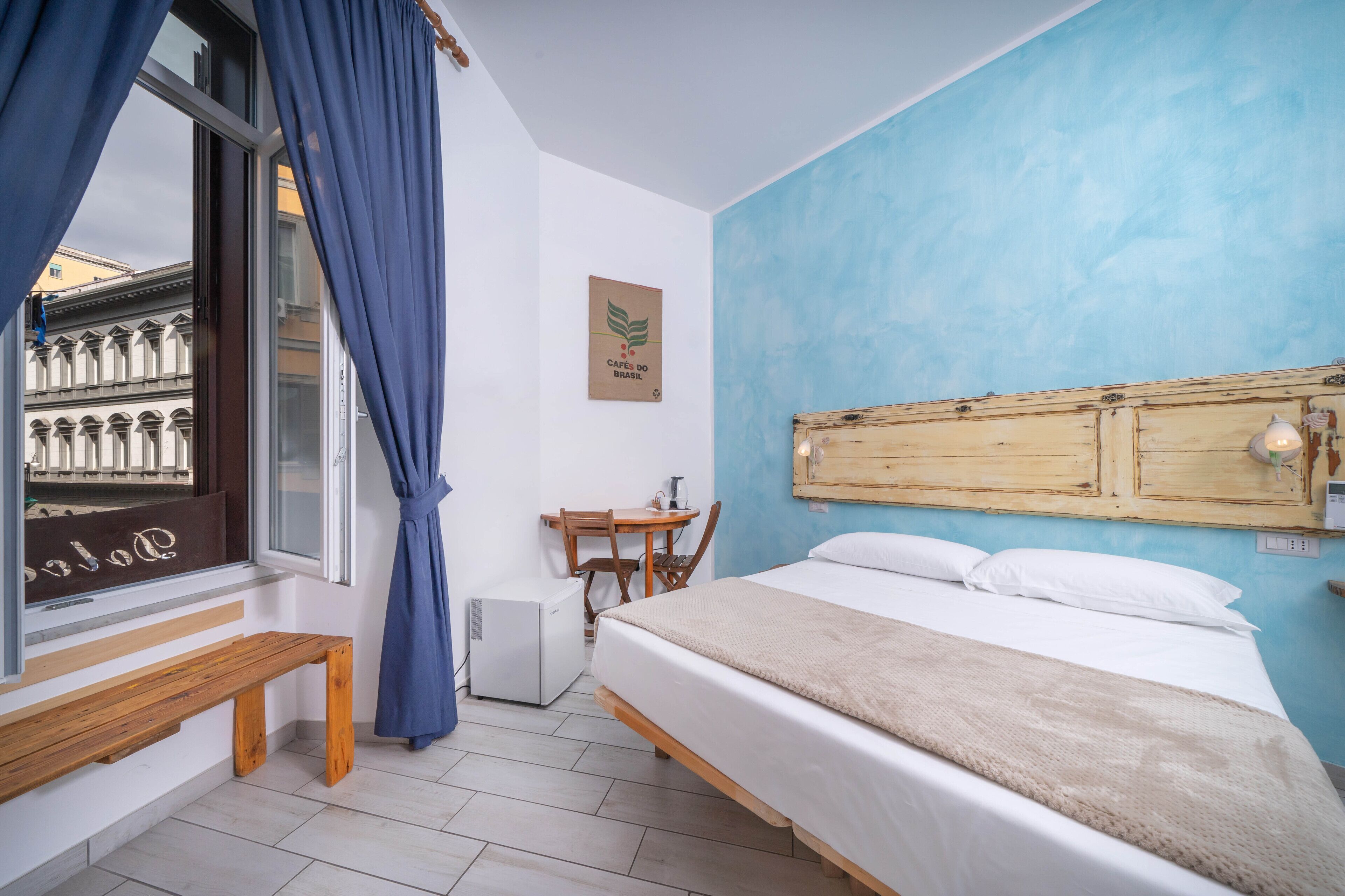 Ein Schlafzimmer mit einem Bett, einem Holz-Kopfteil, einem Fenster mit blauen Vorhängen, einem kleinen Tisch und einer Bank.