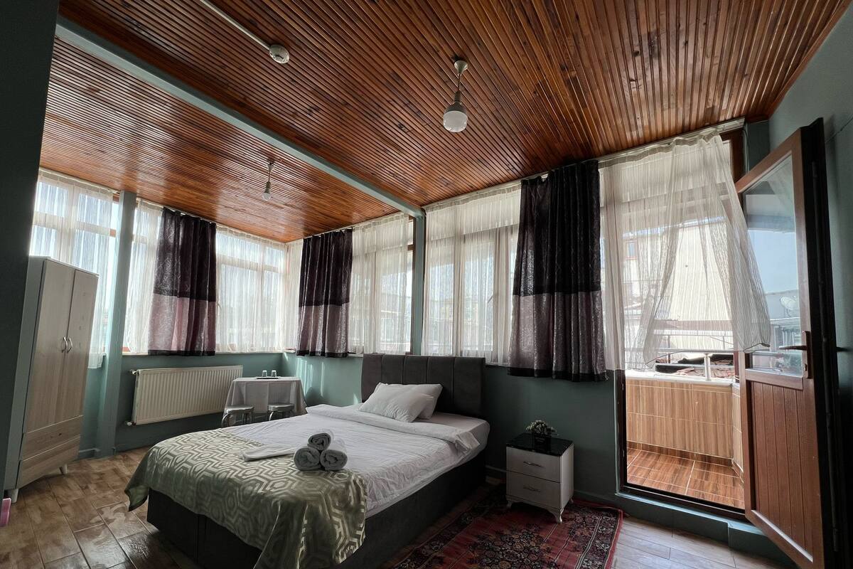 Un dormitorio con techo de madera, una cama con colcha estampada, una mesita de noche, una pequeña mesa con vasos y una ventana con cortinas traslúcidas.