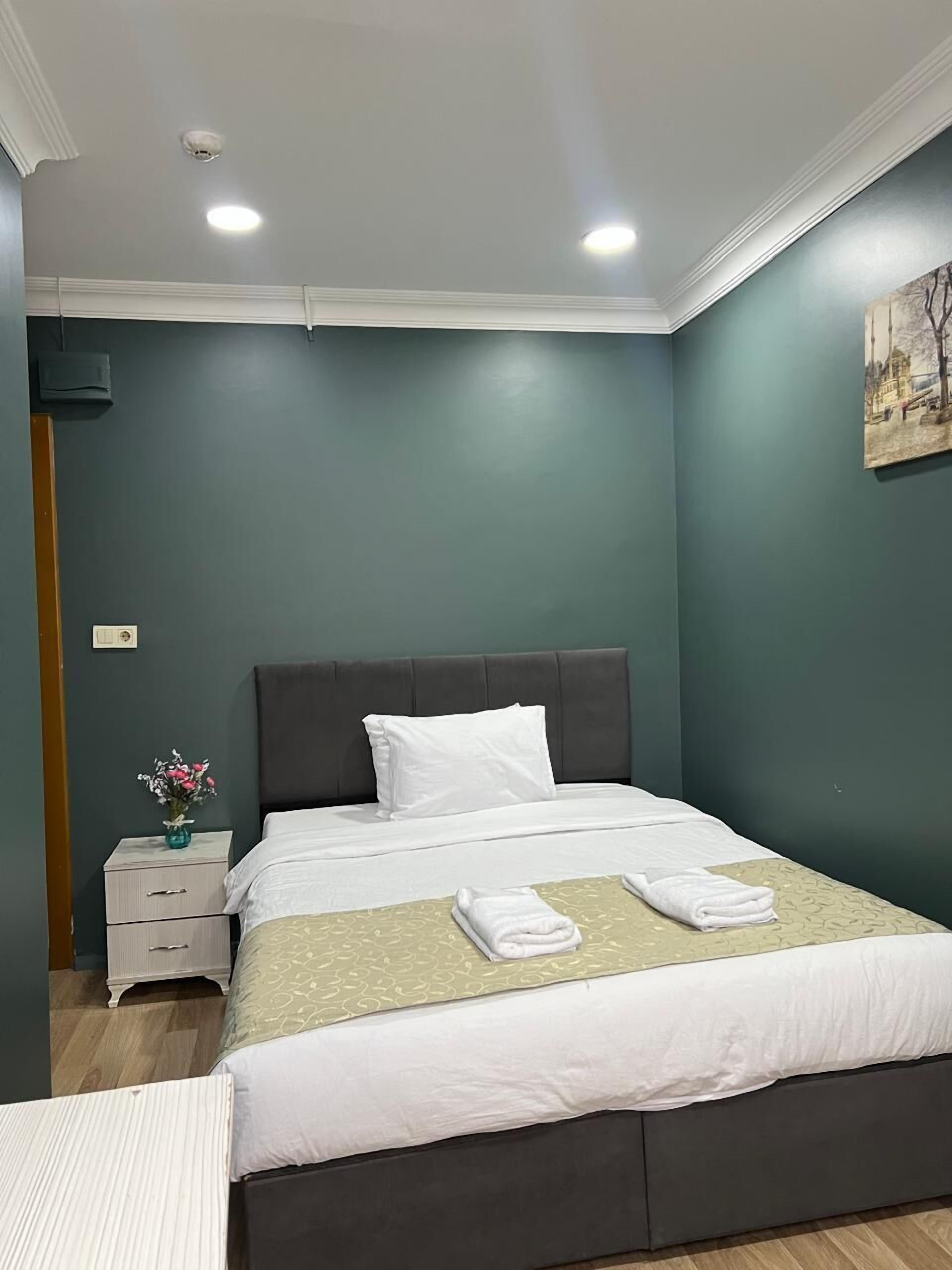 A neatly made bed with white bedding, a dark headboard, and a green patterned bedspread. There is a small bedside table with a vase of flowers and a lamp. The room has teal walls and a ceiling light.