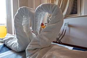 A bed with a white blanket folded into a heart shape, a small wooden table with a glass and a red flower, and a window with blinds. - Vistazzurra B&B (Ancona)