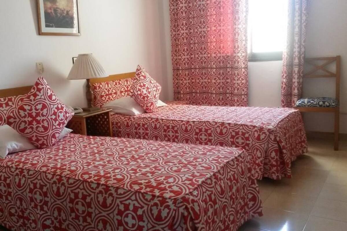 Una habitación con dos camas, cada una con una colcha estampada, una mesita de noche con lámpara y una ventana con cortinas.