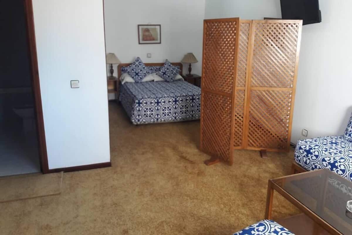 Un dormitorio con cama, mesita de noche, separador de madera, sofá y televisión en la pared.