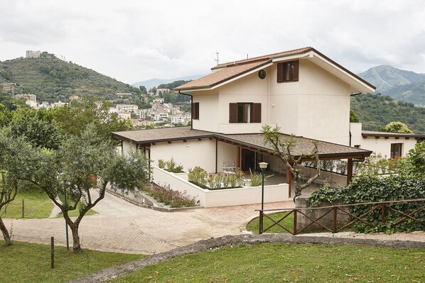 Una casa de dos pisos con balcón, rodeada de vegetación y vistas a una colina con edificios.