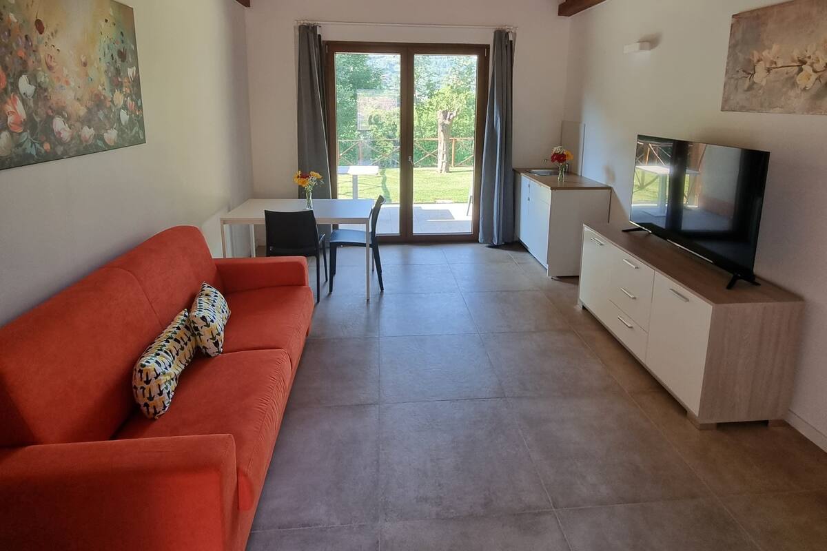 Ein Wohnzimmer mit rotem Sofa, einem Fernseher, einem Essbereich mit Tisch und Stühlen und Blick durch die Glastür.