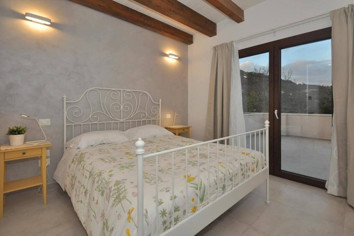 Ein Schlafzimmer mit Bett, Nachttischen, einer Topfpflanze und Blick ins Freie durch eine Schiebetür.