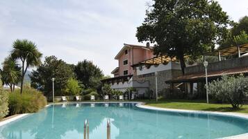 Una piscina con borde curvo, rodeada de un camino de ladrillo. Hay sillas de descanso y un edificio con tejado de azulejos al fondo.