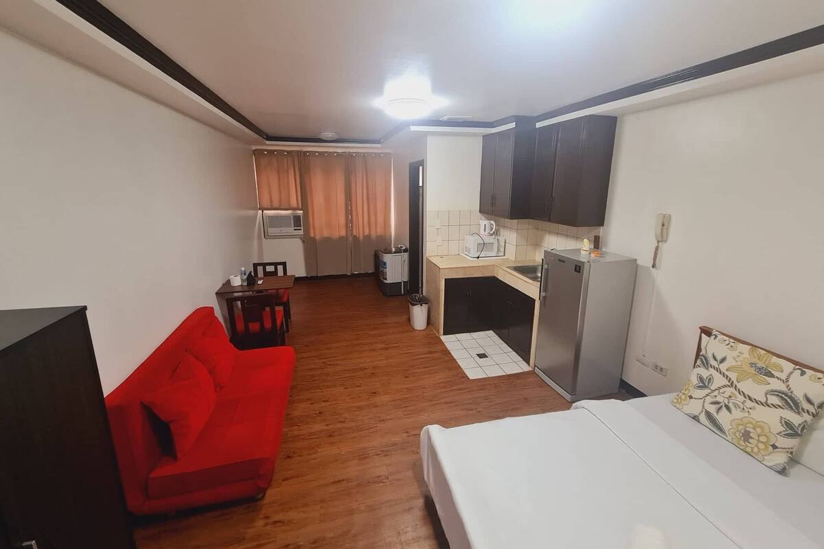 A compact hotel room with a single bed, a red sofa, a kitchenette with a refrigerator, microwave, and sink, and a small dining area with a table and chairs.