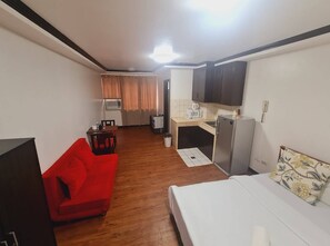 A compact hotel room with a single bed, a red sofa, a kitchenette with a refrigerator, microwave, and sink, and a small dining area with a table and chairs. - Bryan Condo Makati (Makati)