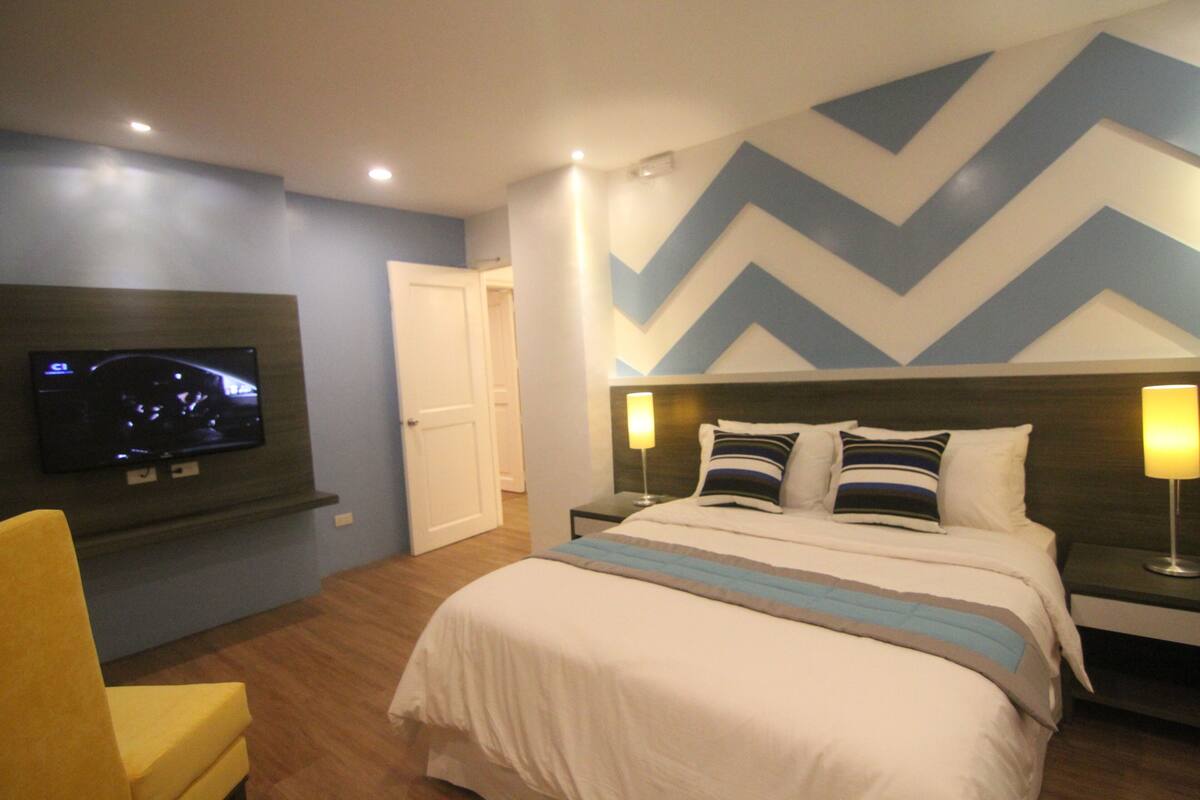A hotel room with a bed, a TV mounted on the wall, a chair, and a door.