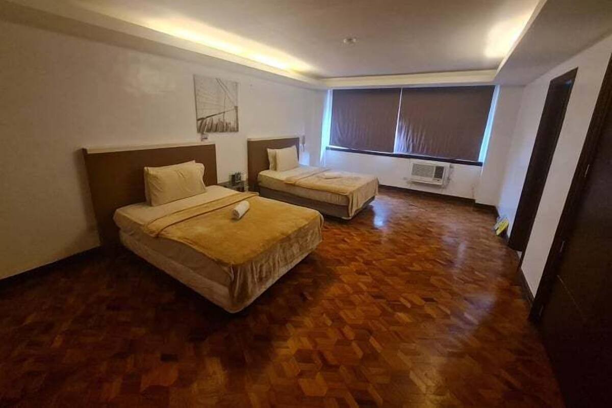 A hotel room with two beds, wooden flooring, a large window with blinds, and an air conditioning unit.