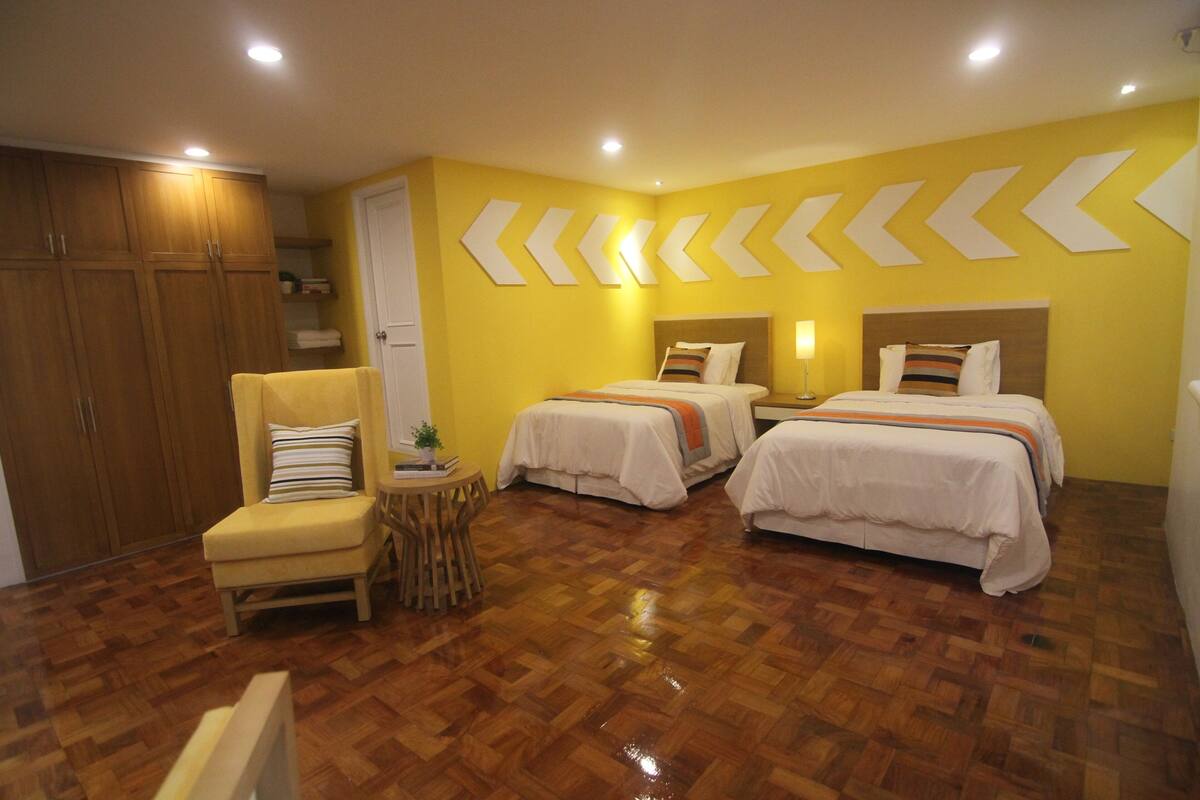 A hotel room with two beds, a chair, a small table with a plant, and wooden flooring.