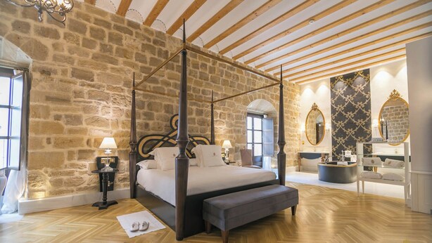 Quarto com cama de dossel, paredes de pedra, vigas de madeira, penteadeira com espelho e balcão de recepção.