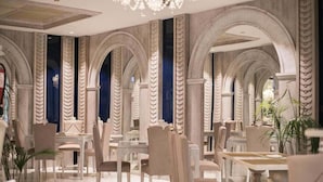 Um hall de hotel espaçoso, com pé direito alto, colunas em arco e um candelabro. Mesas e cadeiras dispostas para os hóspedes.