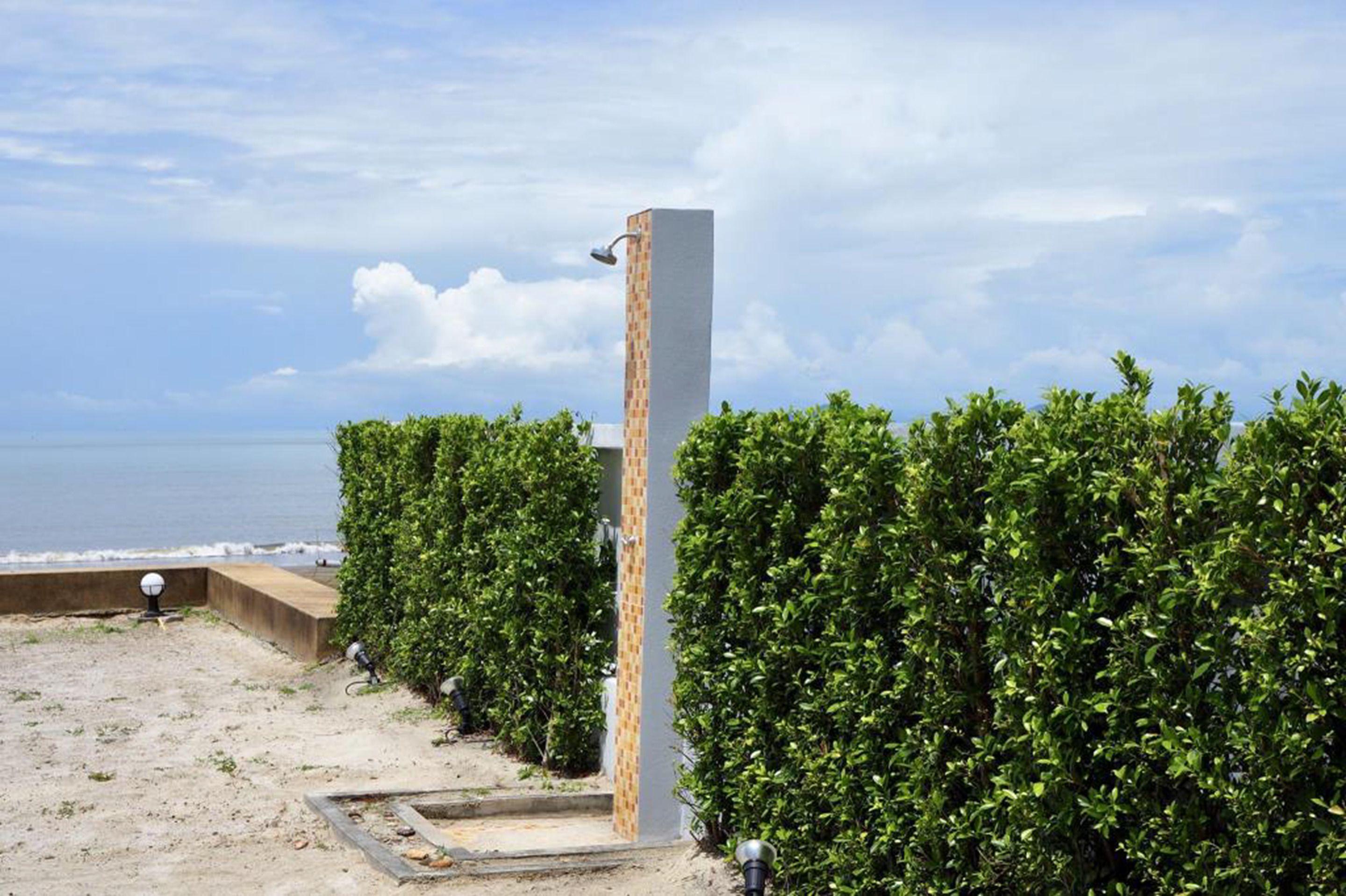 Ein moderner Außenbereich mit Dusche, mit einer hohen, rechteckigen Struktur, umgeben von dichtem Grün, mit Blick auf einen Strand.