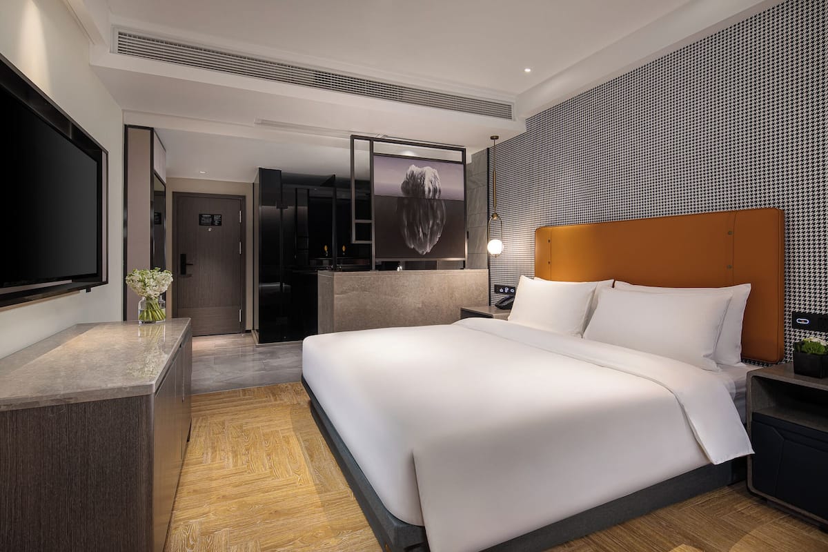 Una habitación de hotel moderna con una cama grande, un televisor de pantalla plana, un minibar y un baño con puerta de vidrio.