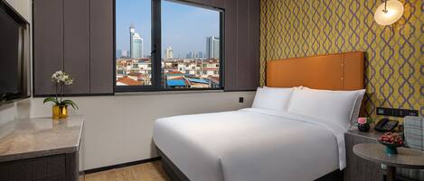 Ein Hotelzimmer mit einem großen Bett, Blick auf die Stadtlandschaft, einem kleinen Tisch mit Blumenvase und einem Schreibtisch mit Telefon.