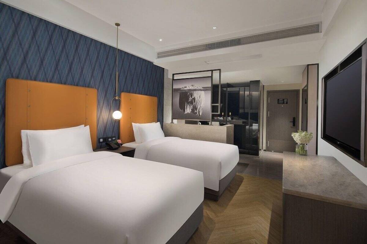 Habitación de hotel con dos camas, un escritorio, televisión y baño visible a través de un panel de vidrio.