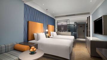 Una habitación de hotel moderna con una cama grande, un sofá, una pequeña mesa redonda con un cuenco de fruta, una televisión y un baño visible a través de una puerta de vidrio.