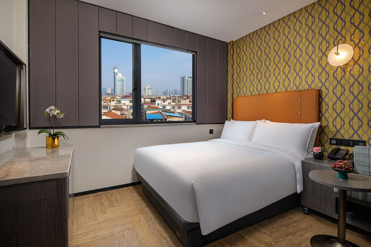 Habitación de hotel con una ventana grande, una cama, una mesita con un florero y un teléfono en la pared.
