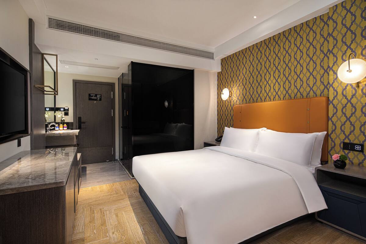 Una habitación de hotel con una cama grande, una mesita de noche con lámpara, un espejo, un baño con lavamanos y artículos de aseo, y una pared con diseño.