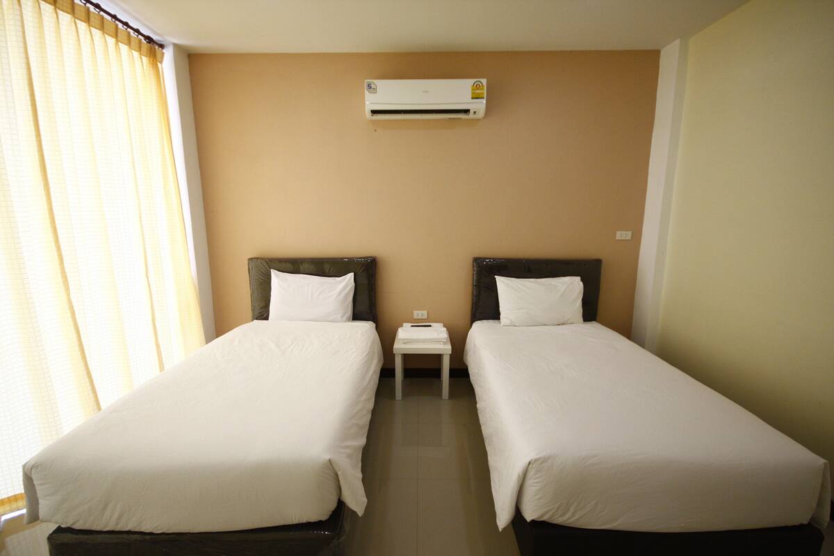 A hotel room with two single beds, a small bedside table, an air conditioning unit on the wall, and a window with curtains.