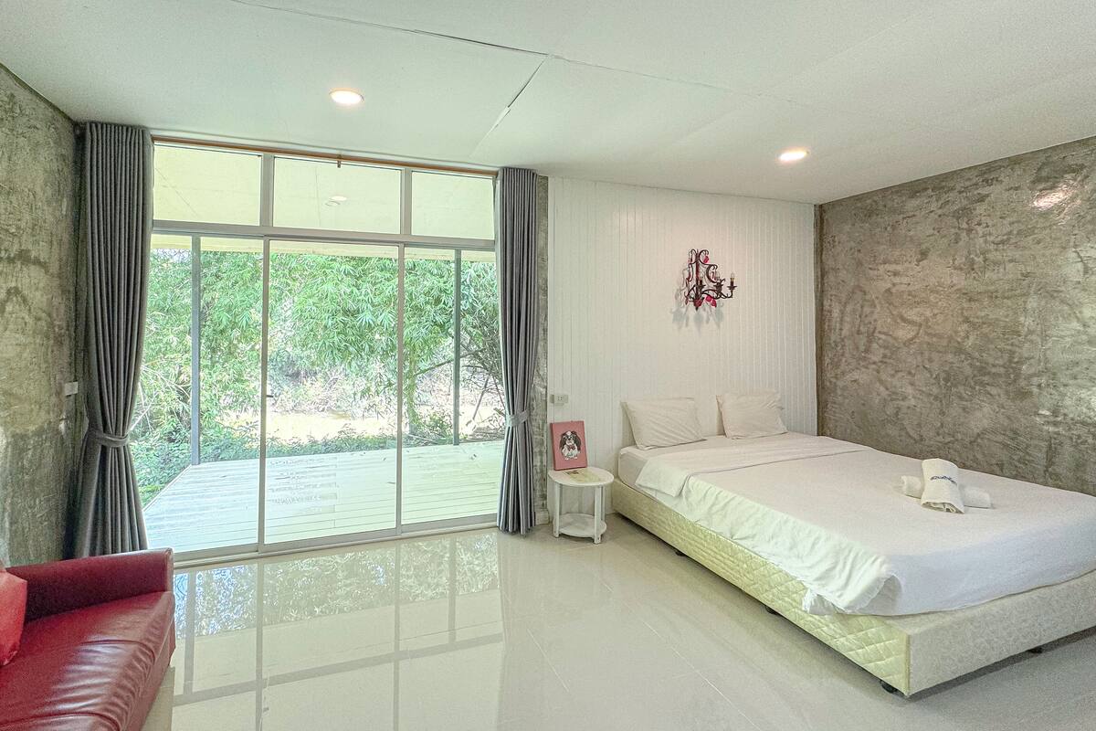A modern bedroom with a large bed, a red sofa, and a view of greenery through the window.