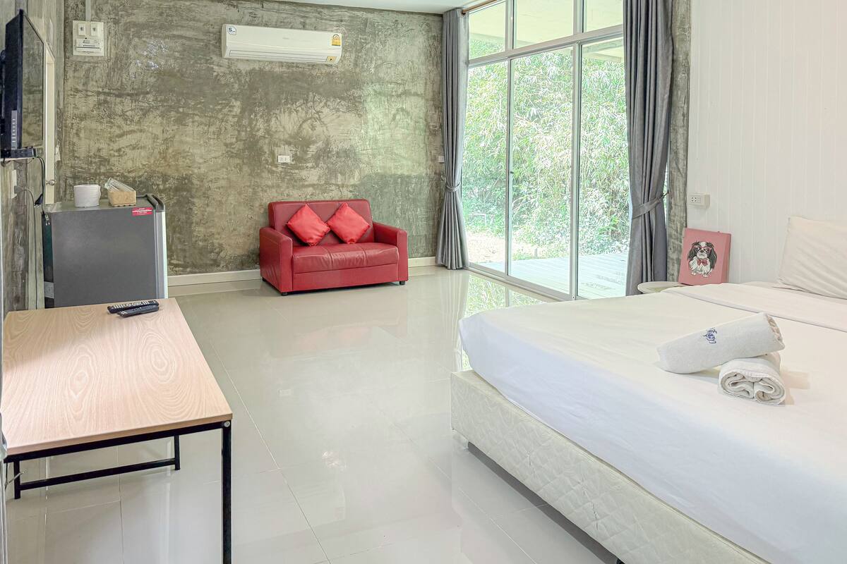 A hotel room with a bed, a red sofa, a small table, and a window with a view of greenery.