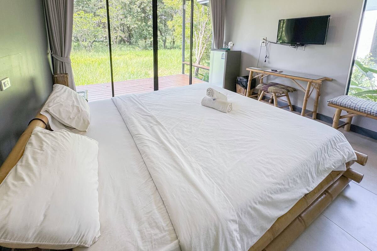 A large bed with white linens, a small desk with a chair, a television on the wall, and a view of greenery outside.