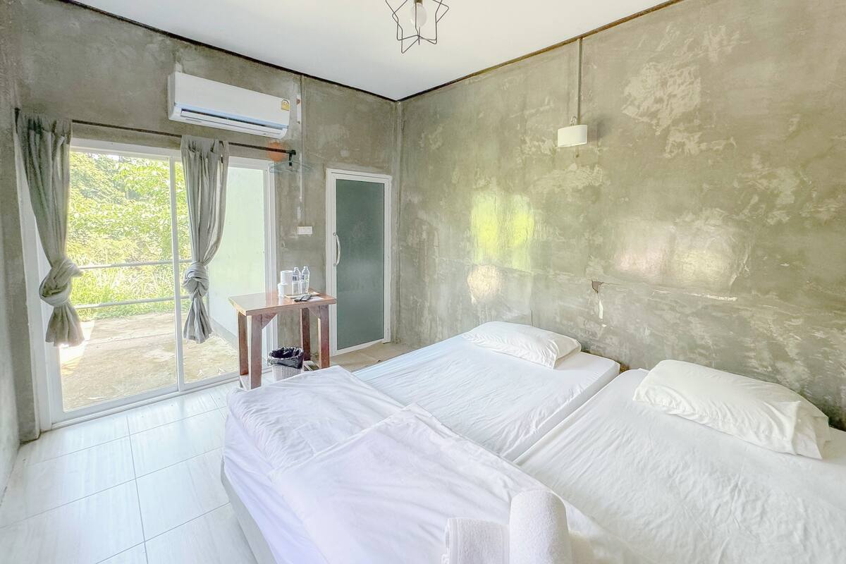 A hotel room with a large bed, a small table with a bottle and glasses, a trash bin, and a sliding door to a balcony.