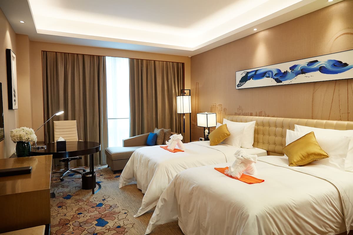 Une chambre d’hôtel avec deux lits, un bureau, une chaise, une lampe et une grande fenêtre avec des rideaux.