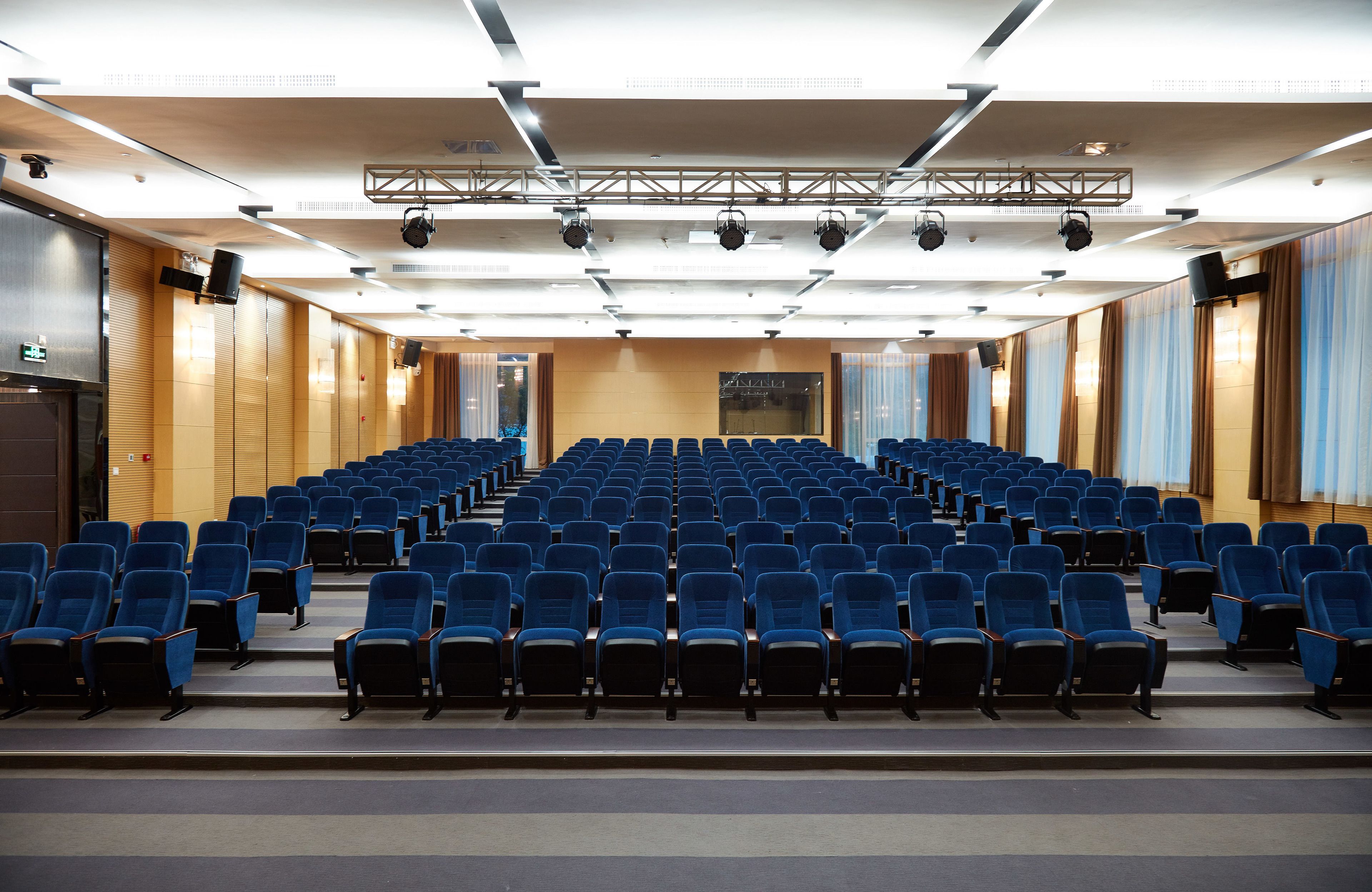 A conference room with rows of blue chairs, a stage with lighting, and a large screen.