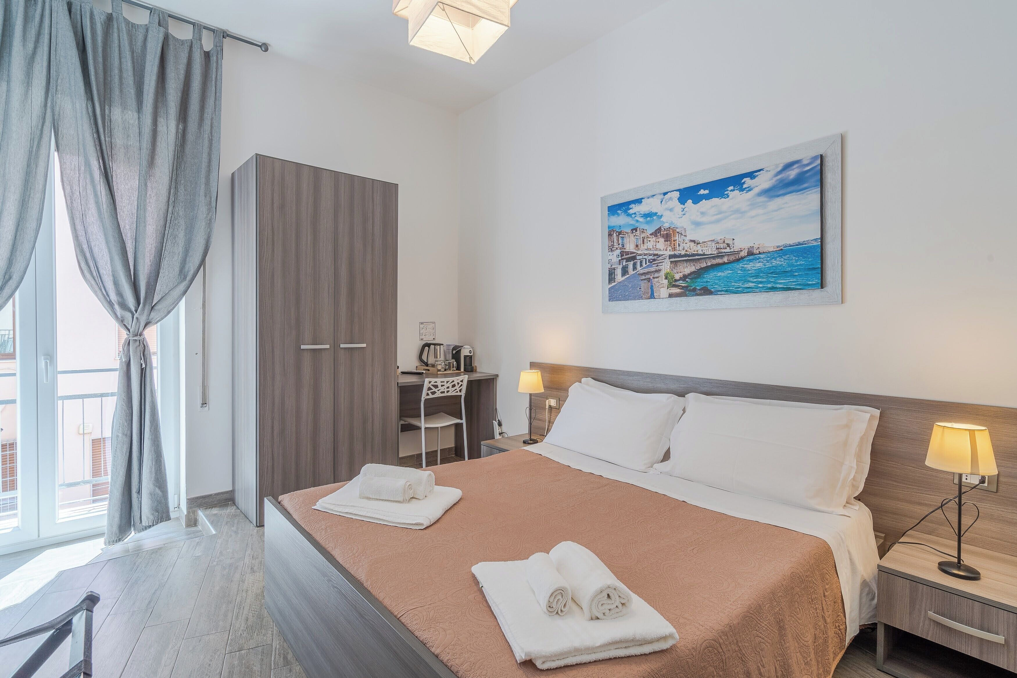 Quarto de hotel com cama, escrivaninha, cadeira, luminária e quadro na parede.
