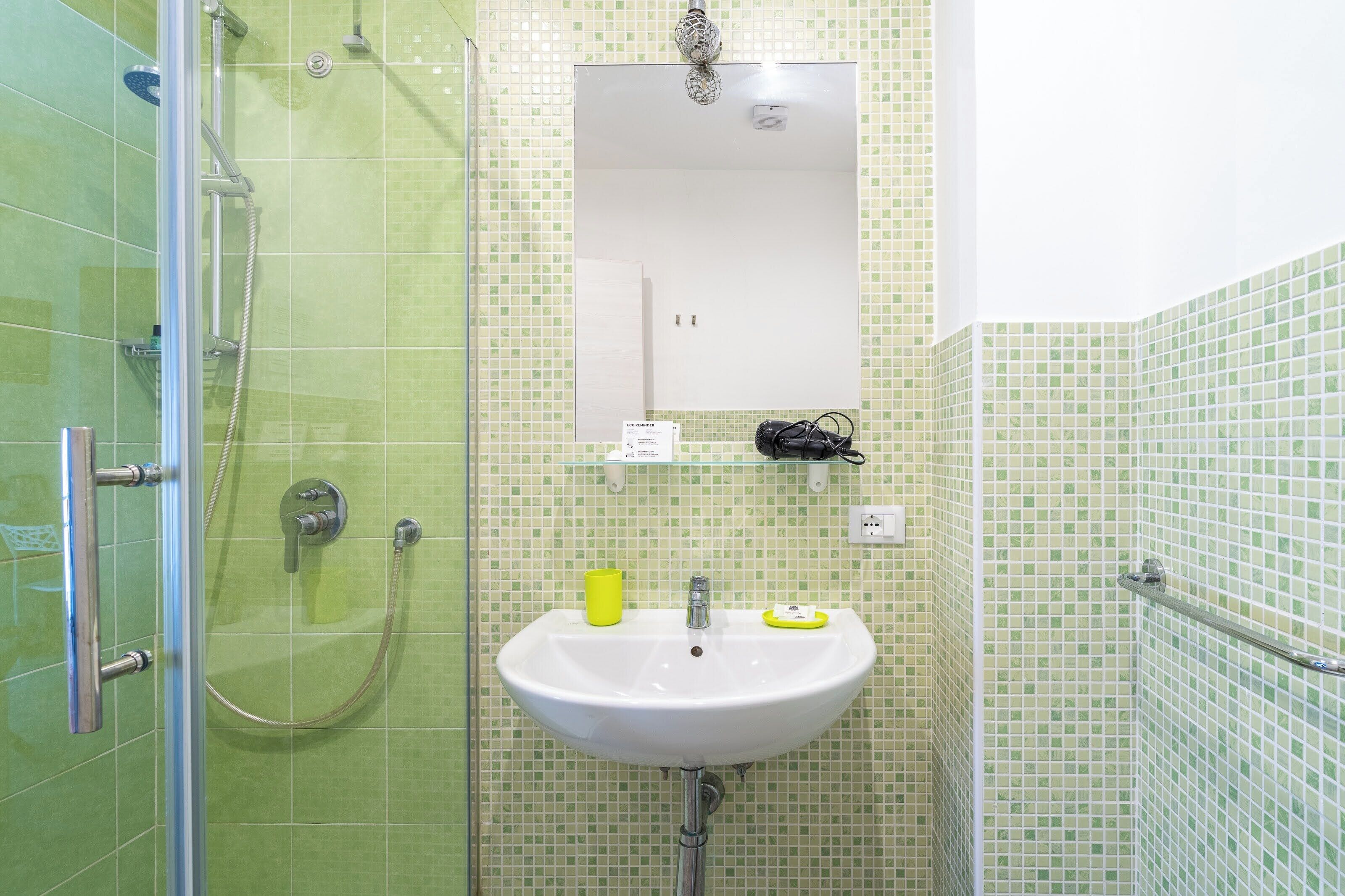 A bathroom with a glass shower enclosure, a white sink with a mirror above it, and a tiled wall with a green pattern.