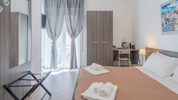 A hotel room with a bed, a desk, a chair, a wardrobe, and a large window with curtains. - Cavour Rooms (Syracuse)