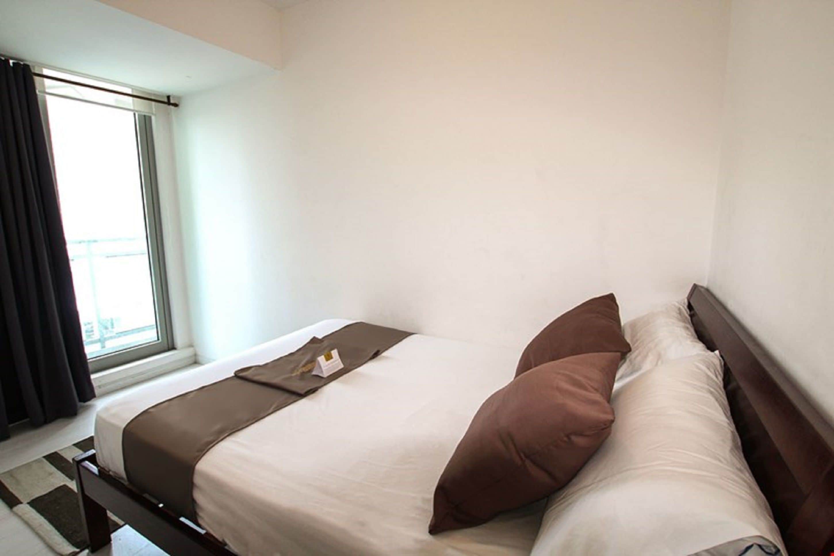 A neatly made bed with a brown bedspread and pillows, a window with curtains, and a view of a balcony.