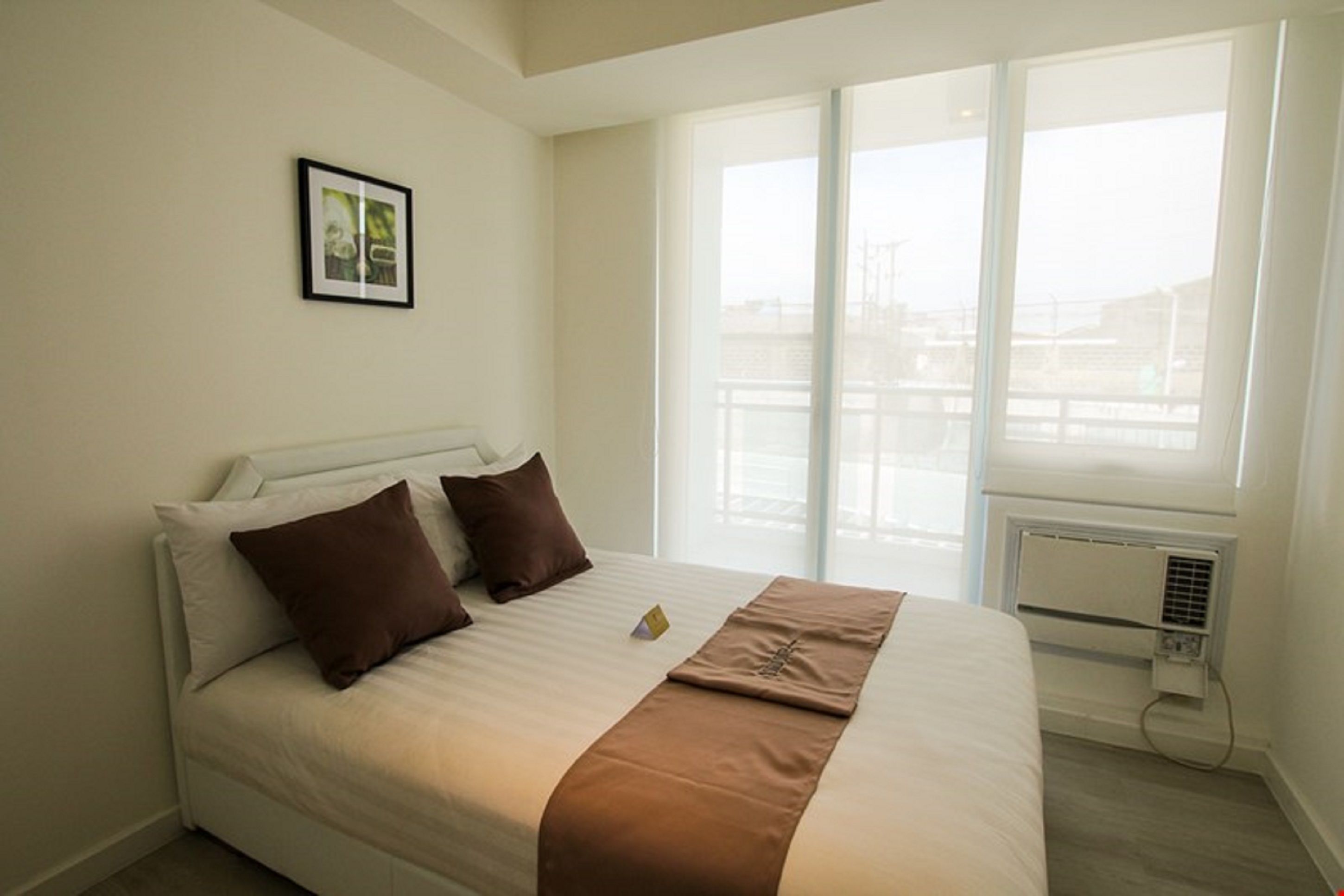 A hotel room with a large bed, white walls, a framed picture on the wall, and a view of a bridge through the window.