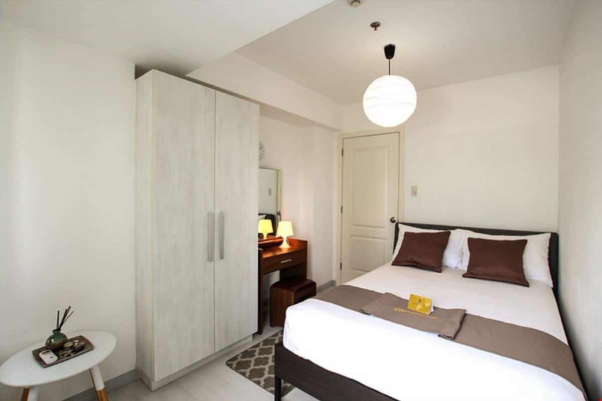 A hotel room with a bed, bedside table, wardrobe, desk, lamp, and a hanging light fixture.