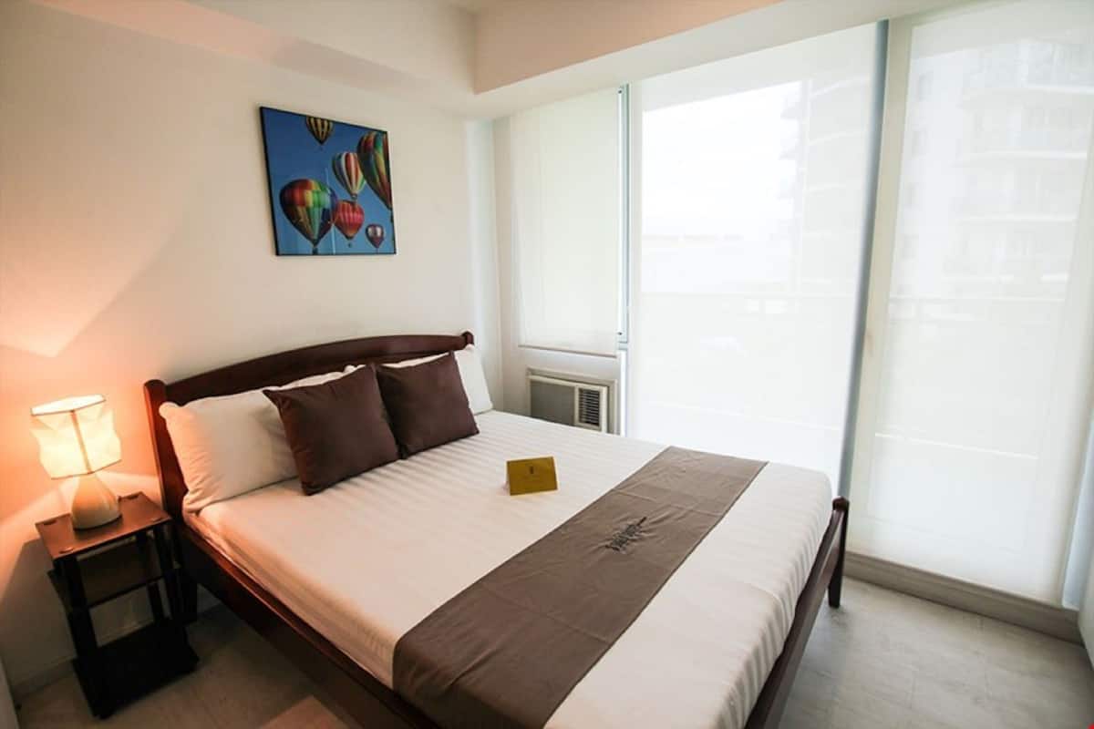 A hotel room with a large bed, a nightstand with a lamp, a painting on the wall, and a window with a view of buildings.