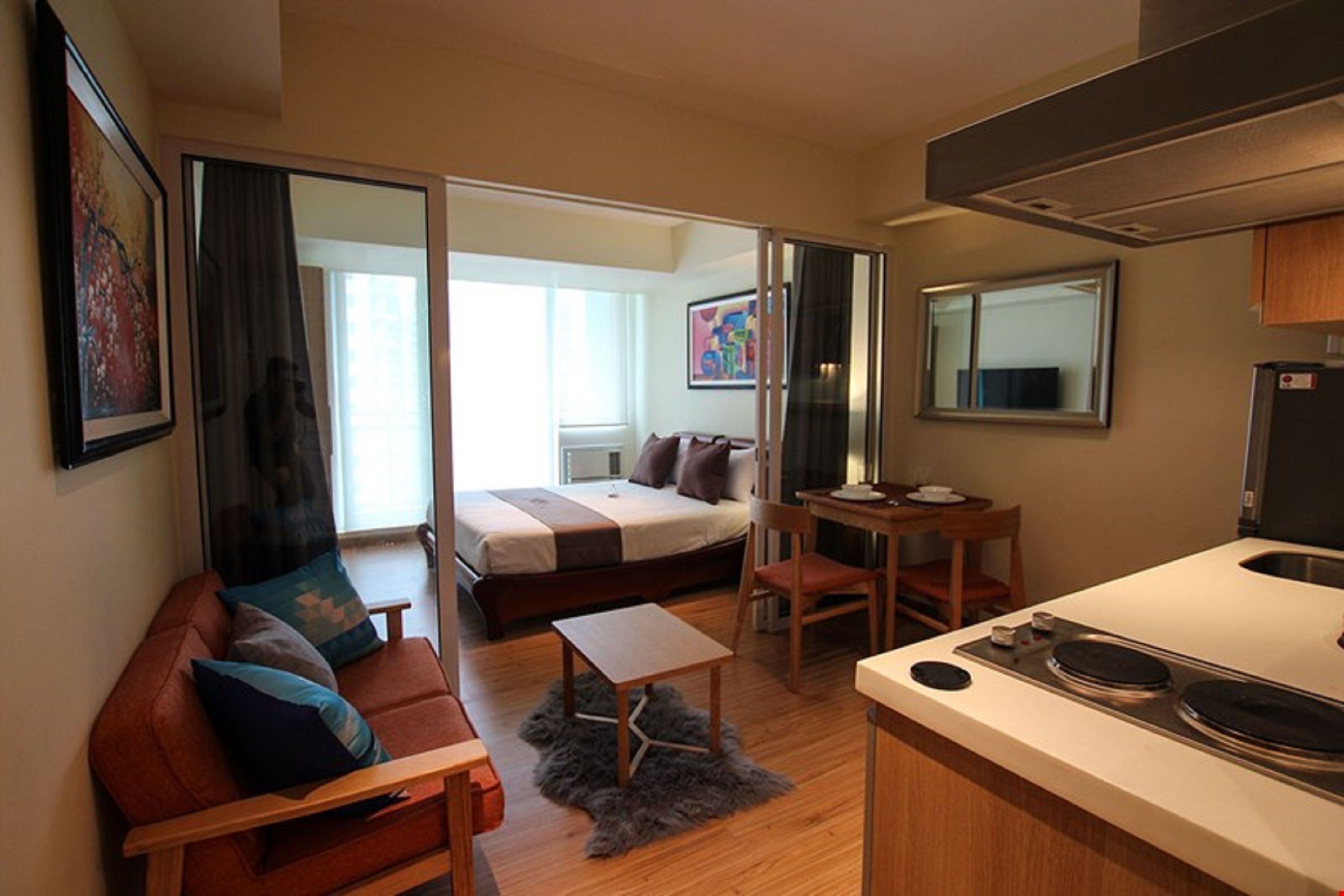 Habitación de hotel compacta con cama, sofá, una mesita, kitchenette y un baño visible a través de una puerta corredera.